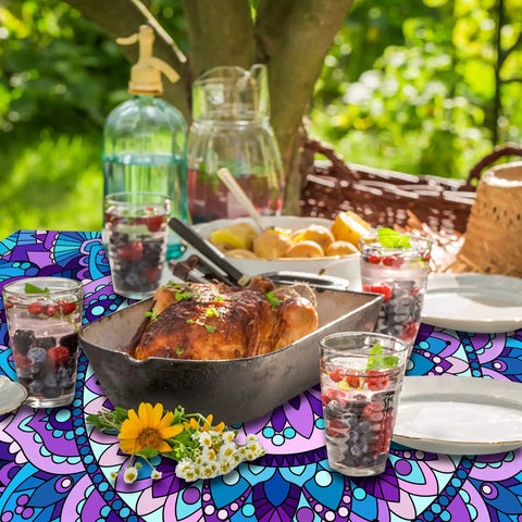 Colorful picnic table setting with food and drinks on a fitted table cover, perfect for outdoor dining under a tree.
