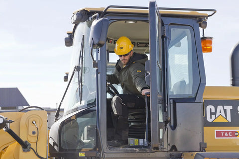Construction worker operating heavy machinery with glass cabin, wearing yellow hard hat and safety gear at a work site.