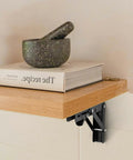 Rugged Matte Black Folding Shelf Brackets, 12 Inch Wall Mounted Hardware For Garage And Workshop on a wooden shelf with a book and bowl.