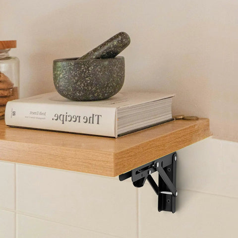 Rugged Matte Black Folding Shelf Brackets, 12 Inch Wall Mounted Hardware For Garage And Workshop on a wooden shelf with a book and bowl.