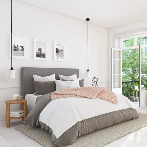 Bright bedroom with grey bed, white linens, pink throw, pendant lights, nightstand, framed art, and open window to greenery