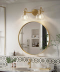 Two-Light Gold Bathroom Vanity Light Fixture With Clear Globe Shades over round mirror in modern bathroom setup