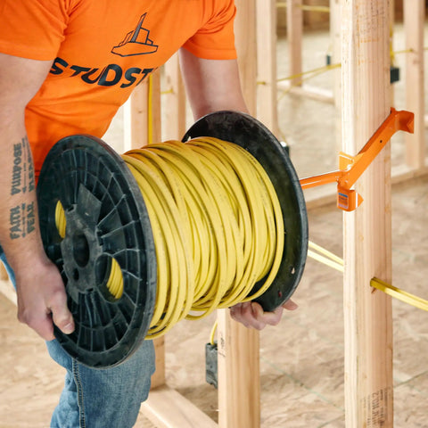 Heavy Duty Wire Dispenser For Stud Framing Mounts To 2x4 And 2x6 Wood Studs With Solid Steel Construction Delivers Fast Load No Fuss Payout For Reels Coils And Cables Made In USA, mounted on wooden studs with a yellow cable spool.