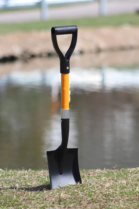 Medium Square Shovel With D Cup Handle 27 Inch Length Durable Build Comfortable Grip For Gardening Landscaping Snow Removal And Light Construction Tools 1 Pack Workhorse Shovel standing upright on grass by water.