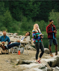 Outdoor fishing scene with four people by a rocky lakeshore, fishing rods in hand and a campfire nearby.