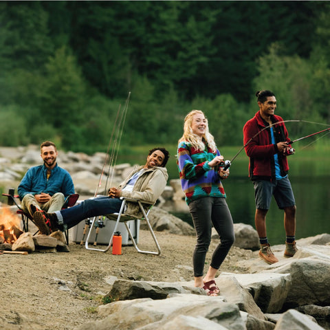 Outdoor fishing scene with four people by a rocky lakeshore, fishing rods in hand and a campfire nearby.