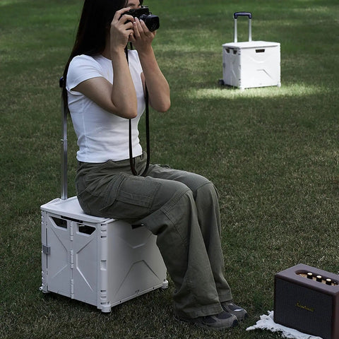 Person sitting on a collapsible rolling crate on wheels for teachers with heavy duty plastic tote basket in a grassy area.