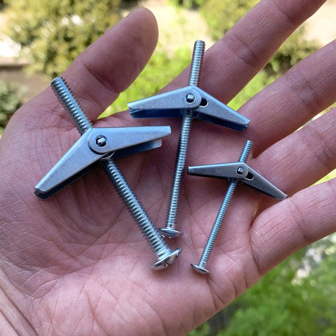 Hand holding three toggle bolts from Toggle Bolt Kit 29-Piece Zinc Plated Steel Set With One-Eighth Inch, Three-Sixteenth Inch, And One-Fourth Inch Toggle Bolts Plus Wing Nuts For Hanging Heavy Stuff On Drywall.