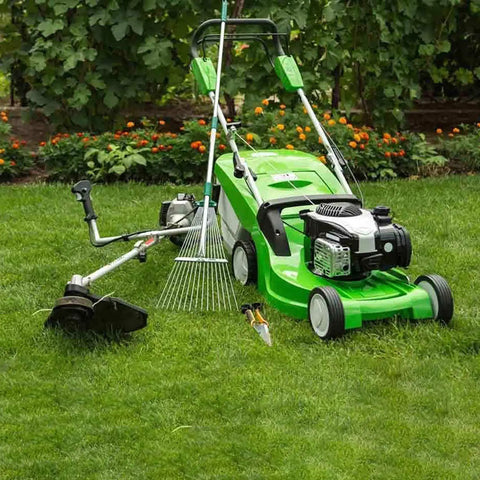 Green lawn mower and weed trimmer on lush grass with garden tools and flowers in background.