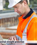 Construction worker in orange safety vest and white helmet using a laptop on a building site.