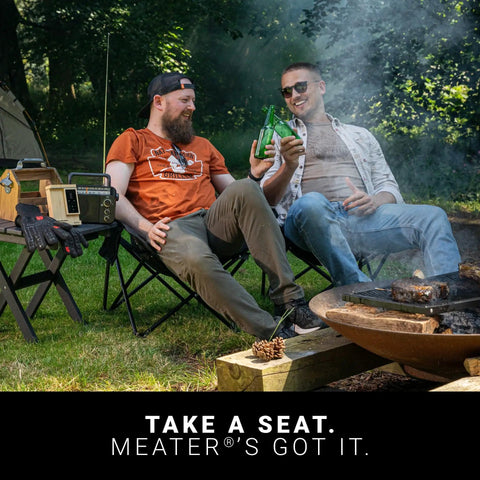 Two men sitting by a campfire, enjoying drinks and relaxing outdoors with a Four Probe Wireless Smart Meat Thermometer nearby.