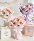 Bouquets of pink and purple carnations in pastel wrapping with Happy Mother's Day gifts and cupcakes on a decorated table.