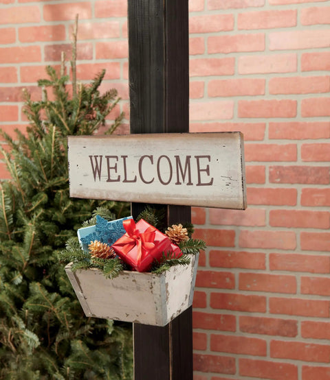 Welcome sign with pine decoration and gift box on post by brick wall.