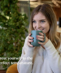Woman smiling while holding a Slick App Controlled 14 Oz Smart Heated Coffee Mug With Rechargeable Base And Glow Led Display.