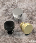 Three knurled round cabinet knobs in brushed satin nickel, black, and gold on a textured surface.