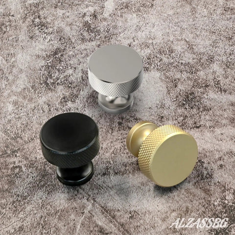 Three knurled round cabinet knobs in brushed satin nickel, black, and gold on a textured surface.