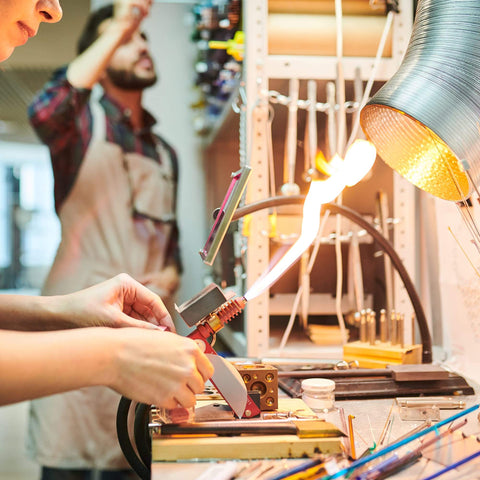 Person using tools in workshop with bright flame, wearing apron amidst organized shelves. Industrial setting with focus on precise craftsmanship.