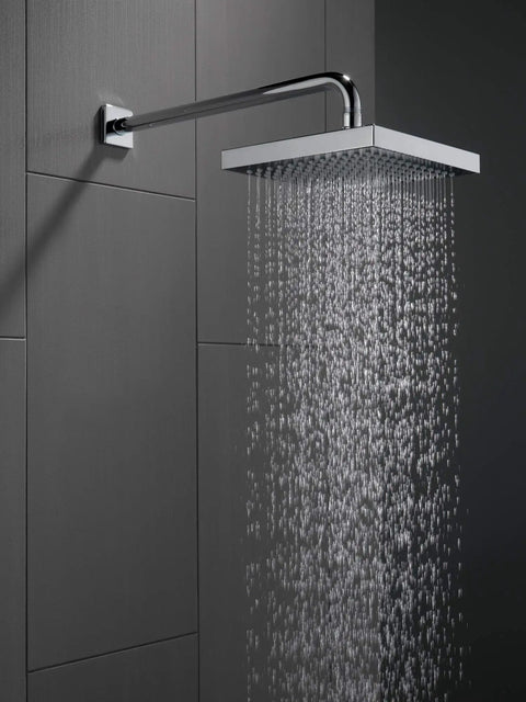 16 Inch Black Stainless Shower Arm Trim With One Half IPS Threads, Wall Mount Design For A Fresh Shower Upgrade, Durable Finish That Lasts, Flange And Shower Head Sold Separately, attached to a tile wall with water flowing.