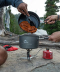 Camping scene with two people cooking using a portable stove; a pot of stew is being prepared outdoors.