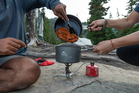 Camping scene with two people cooking using a portable stove; a pot of stew is being prepared outdoors.
