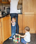 Two toddlers playing with blocks in a kitchen, woman reaching into a cupboard above, wooden cabinets with silver handles visible.