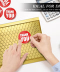 Hands placing a thank you sticker on a Gold Bubble Mailers 6x10 For Jewelry And Makeup Supplies 50 Pack Metallic Envelopes With Strong Self Adhesive Seal Waterproof Padding Durable Bulk Mailers That Look Luxe And Ship Safe on a desk.