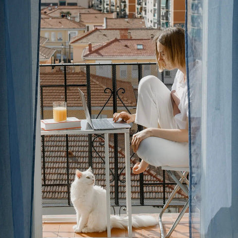 Woman working on balcony with laptop, books, and juice, with Pet Balcony Net For Cats And Dogs 16.5 Ft By 2.5 Ft Anti Fall Mesh Screen.