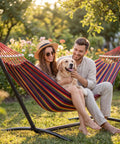 Double Hammock With Heavy Duty Steel Stand Portable Easy Assembly 600Lb Weight Capacity Adjustable Hooks Cotton Comfort Durable Frame Carrying Bag Included Ideal For Indoor Yard Camping And Outdoor Rainbow with couple and dog in garden setting