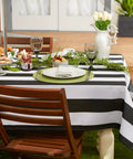 Cabana Stripe Outdoor Zippered Tabletop Cover 60 by 84 Inches Stain Resistant and Waterproof Machine Washable Easy Care for Patio Tables That Seat 6 to 8 Black on wooden outdoor table with white dishes and greenery centerpiece.