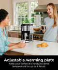 Two women enjoying coffee near a 12 Cup Programmable Coffee Brewer With Classic Or Rich Brew Styles in a kitchen setting.
