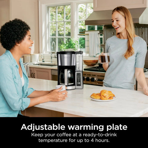 Two women enjoying coffee near a 12 Cup Programmable Coffee Brewer With Classic Or Rich Brew Styles in a kitchen setting.
