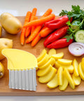 Front view of yellow crinkle cutter with stainless steel ridged blade on wooden board beside potatoes, carrots and peppers.
