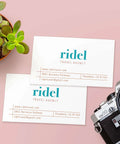 Two white business cards front view with teal ridel logo and orange travel agency text on pink background beside a camera.