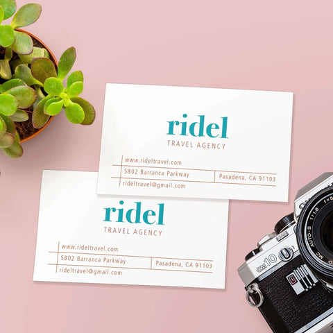 Two white business cards front view with teal ridel logo and orange travel agency text on pink background beside a camera.