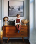 Child in red overalls sitting on a wooden cabinet with decor, under a framed black-and-white photograph on the wall.