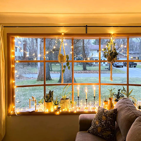 Warm White 300 LED Christmas String Lights With Clear Wire For Indoor Outdoor Use Waterproof Durable Eight Modes Including Sequential And Chase Effects For Steady Glow Plug In Fairy Lights For Christmas Tree Bedroom And Cozy Vibe Decor illuminating a window with plants and candles.