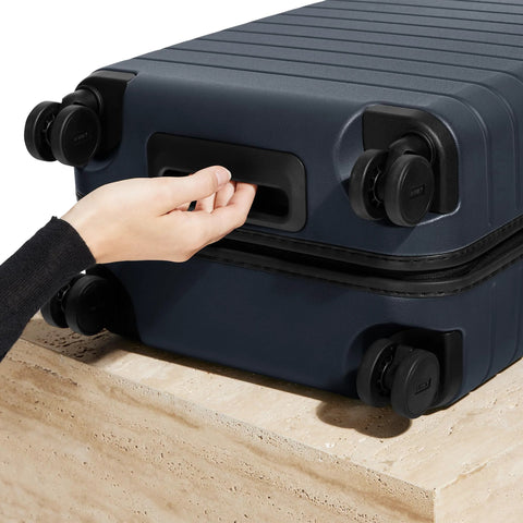 Close-up of navy Bigger Carry On Luggage From Away, featuring spinner wheels and TSA lock, on a stone surface.