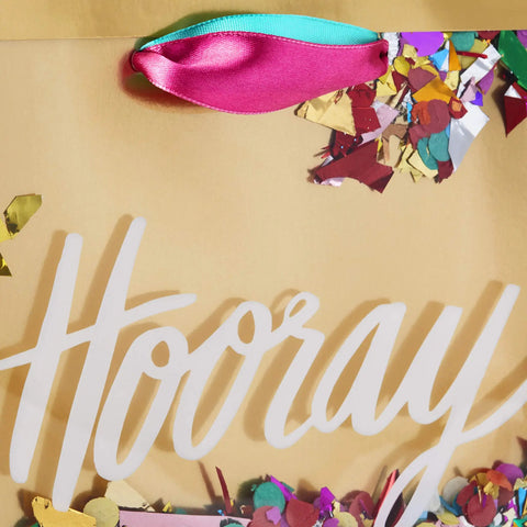 Close-up of a Medium Gift Bag With Tissue Paper And Hooray Confetti Design in gold with pink teal purple ribbon and confetti.