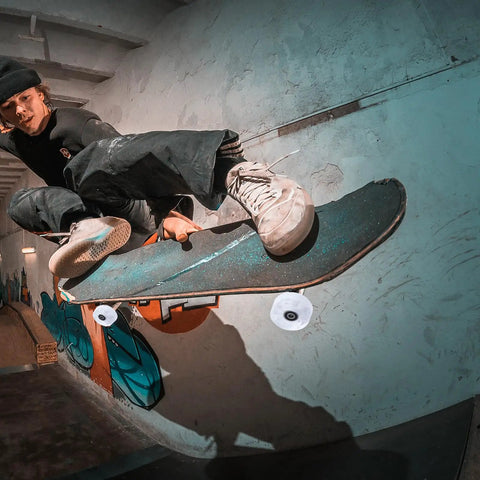 Person performing skateboard trick indoors with graffiti backdrop, showcasing intense jump and dynamic movement.