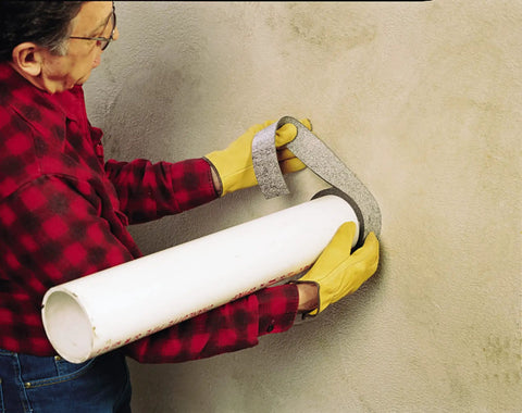 Person wrapping Graphite Based Intumescent Fire Barrier Wrap Strip 2 Inch By 40 Foot Flexible Firestop around pipe in wall.