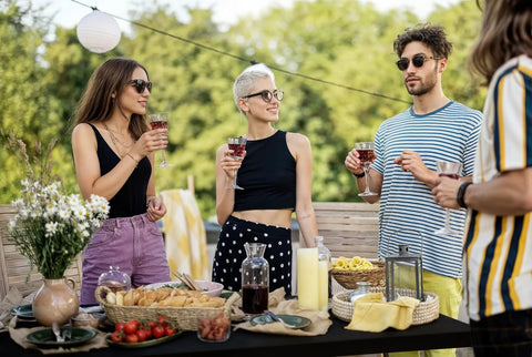 People gathered around a table with food and drinks, outdoors in a garden setting, during a casual gathering.