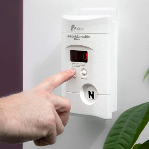 Plug-In Carbon Monoxide Detector With 9V Battery Backup, Digital LED Display, 85 Decibel Alarm on wall with finger pressing test button