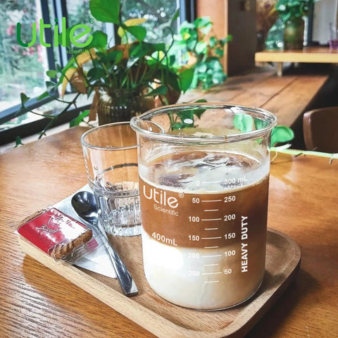 400ml beaker with iced coffee and cream next to a glass and snack on a wooden table by the window