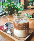 Beaker and glass with coffee on a wooden tray next to plant, focus on spouted beaker with double scaled graduation marks