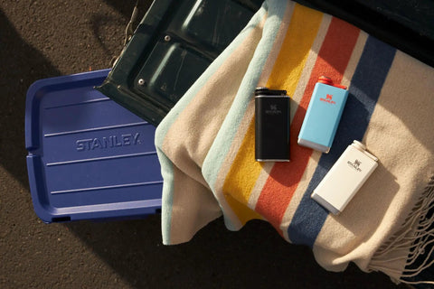 Three hip flasks on a striped blanket next to a blue container, including Pre Party 8 Oz Leakproof Hip Flask With Hinged Cap, Insulated Stainless Steel Liquor Container, BPA Free, Hammertone Green, Perfect On The Go Party Starter For Travelers.