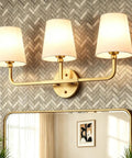 Top Tier Three Light Brushed Gold Vanity Lights With Flared White Fabric Shades Smooth Bathroom Mirror Fixture and modern wall sconces above a mirror.