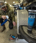 Quicksilver 2-4-C marine grease with PTFE, 8 oz tube, on diamond-plate step; person kneels with a grease gun.