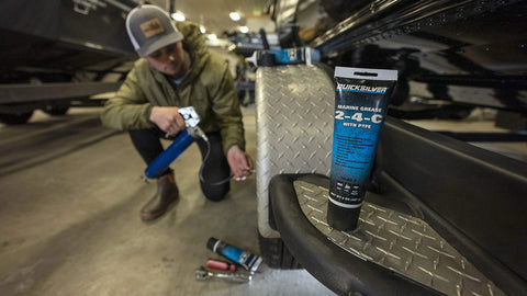 Quicksilver 2-4-C marine grease with PTFE, 8 oz tube, on diamond-plate step; person kneels with a grease gun.