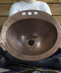 Person holding Bell Drop In Handmade Copper Bath Sink With Four Inch Faucet Holes And Overflow, showcasing antique copper texture.