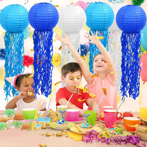 Colorful Mermaid Jellyfish Paper Lanterns Hanging Set For Under The Sea Theme Parties, Ocean Party Decor, Birthday Decorations, Kid Friendly Baby Shower Or Nursery, 6 Pack, Light Not Included; children enjoying a party under blue and white lanterns above a decorated table.